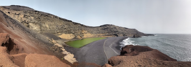 A rugged coastal landscape with a steep rocky cliff and a green lagoon nestled between the rocks. The foreground features reddish-brown volcanic terrain leading down to a dark sandy beach. The sea extends to the horizon, creating a stark contrast with the land.