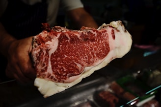 A person holding a large piece of marbled raw beef, showcasing its intricate fat marbling and deep red color. The beef is prominently displayed against a dark background, possibly in a butcher shop setting.