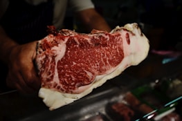 A person holding a large piece of marbled raw beef, showcasing its intricate fat marbling and deep red color. The beef is prominently displayed against a dark background, possibly in a butcher shop setting.