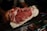 A person holding a large piece of marbled raw beef, showcasing its intricate fat marbling and deep red color. The beef is prominently displayed against a dark background, possibly in a butcher shop setting.