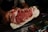 A person holding a large piece of marbled raw beef, showcasing its intricate fat marbling and deep red color. The beef is prominently displayed against a dark background, possibly in a butcher shop setting.