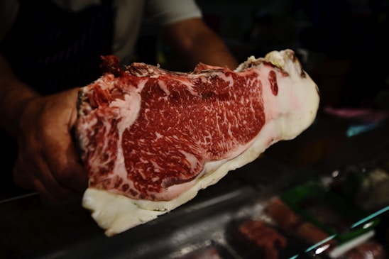 A person holding a large piece of marbled raw beef, showcasing its intricate fat marbling and deep red color. The beef is prominently displayed against a dark background, possibly in a butcher shop setting.