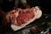 A person holding a large piece of marbled raw beef, showcasing its intricate fat marbling and deep red color. The beef is prominently displayed against a dark background, possibly in a butcher shop setting.