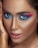 A close-up portrait featuring a person with dramatic, brightly colored eye makeup in shades of pink and blue. The skin has a smooth, flawless appearance with a warm, bronzed tone. The lips are slightly parted with a glossy pink hue and the eyes are wide and captivating.