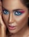 A close-up portrait featuring a person with dramatic, brightly colored eye makeup in shades of pink and blue. The skin has a smooth, flawless appearance with a warm, bronzed tone. The lips are slightly parted with a glossy pink hue and the eyes are wide and captivating.