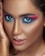 A close-up portrait featuring a person with dramatic, brightly colored eye makeup in shades of pink and blue. The skin has a smooth, flawless appearance with a warm, bronzed tone. The lips are slightly parted with a glossy pink hue and the eyes are wide and captivating.