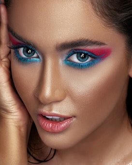 A close-up portrait featuring a person with dramatic, brightly colored eye makeup in shades of pink and blue. The skin has a smooth, flawless appearance with a warm, bronzed tone. The lips are slightly parted with a glossy pink hue and the eyes are wide and captivating.