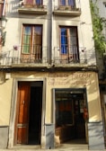 A building facade featuring two stories with wooden-framed windows, each shaded by metal railings and decorated with pots. Below, there is a shop displaying guitars in the glass window and a wooden door to the left. The sign above reads 'Casa Ferrer, Construcción de Guitarras', indicating a guitar shop established in 1879.