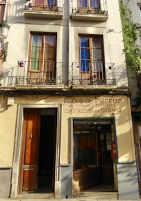 A building facade featuring two stories with wooden-framed windows, each shaded by metal railings and decorated with pots. Below, there is a shop displaying guitars in the glass window and a wooden door to the left. The sign above reads 'Casa Ferrer, Construcción de Guitarras', indicating a guitar shop established in 1879.