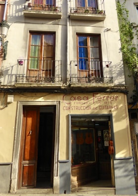 A building facade featuring two stories with wooden-framed windows, each shaded by metal railings and decorated with pots. Below, there is a shop displaying guitars in the glass window and a wooden door to the left. The sign above reads 'Casa Ferrer, Construcción de Guitarras', indicating a guitar shop established in 1879.