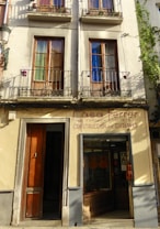 A building facade featuring two stories with wooden-framed windows, each shaded by metal railings and decorated with pots. Below, there is a shop displaying guitars in the glass window and a wooden door to the left. The sign above reads 'Casa Ferrer, Construcci&oacute;n de Guitarras', indicating a guitar shop established in 1879.