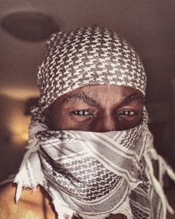 A person is wearing a patterned scarf that covers their head and lower face, leaving only their eyes visible. The scarf features a traditional design with a mix of dark and light tones. The person is gazing directly at the camera, creating a strong visual focus on their eyes.