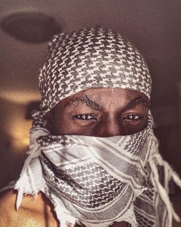 A person is wearing a patterned scarf that covers their head and lower face, leaving only their eyes visible. The scarf features a traditional design with a mix of dark and light tones. The person is gazing directly at the camera, creating a strong visual focus on their eyes.