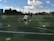 A child is playing soccer on an outdoor field, running towards a ball between two rows of colored cones. The coach stands in the background, observing the child. The field is lush and green, with trees and buildings visible in the distance. The sky is partly cloudy, indicating a sunny day.