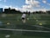 A child is playing soccer on an outdoor field, running towards a ball between two rows of colored cones. The coach stands in the background, observing the child. The field is lush and green, with trees and buildings visible in the distance. The sky is partly cloudy, indicating a sunny day.