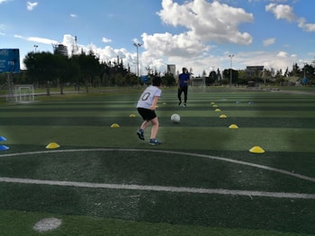 A child is playing soccer on an outdoor field, running towards a ball between two rows of colored cones. The coach stands in the background, observing the child. The field is lush and green, with trees and buildings visible in the distance. The sky is partly cloudy, indicating a sunny day.