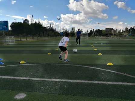 A child is playing soccer on an outdoor field, running towards a ball between two rows of colored cones. The coach stands in the background, observing the child. The field is lush and green, with trees and buildings visible in the distance. The sky is partly cloudy, indicating a sunny day.
