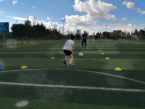 A child is playing soccer on an outdoor field, running towards a ball between two rows of colored cones. The coach stands in the background, observing the child. The field is lush and green, with trees and buildings visible in the distance. The sky is partly cloudy, indicating a sunny day.