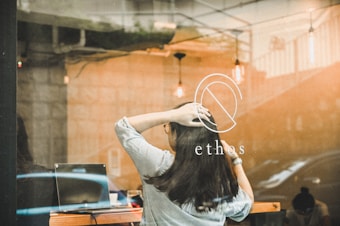 A woman with long hair is seen through a window with her hands on her head, sitting at a wooden table. The window has the word 'ethos' and a logo printed on it. There is a laptop on the table, along with some small items. The reflection on the glass shows hints of an urban environment. The interior is warmly lit with hanging light bulbs visible and elements of industrial design.