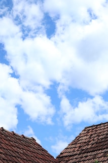 Two rooftops with terracotta tiles are visible against a partly cloudy sky, with fluffy white clouds dispersed across a bright blue background.