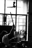 A person is reaching up with a long stick towards a ceiling fan inside a room. The room has an open window with grilles, through which light is streaming in. Various items are seen hanging on a drying rack near the window.
