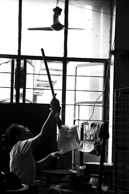 A person is reaching up with a long stick towards a ceiling fan inside a room. The room has an open window with grilles, through which light is streaming in. Various items are seen hanging on a drying rack near the window.