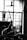 A person is reaching up with a long stick towards a ceiling fan inside a room. The room has an open window with grilles, through which light is streaming in. Various items are seen hanging on a drying rack near the window.