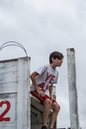 A young boy is crouched on a weathered, white-painted wooden platform, appearing to prepare to jump or dive. He is wearing a white T-shirt with large red letters spelling 'YEAH' and red shorts. The backdrop is a cloudy sky, giving an overcast atmosphere.