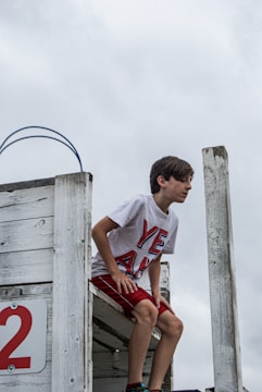 A young boy is crouched on a weathered, white-painted wooden platform, appearing to prepare to jump or dive. He is wearing a white T-shirt with large red letters spelling 'YEAH' and red shorts. The backdrop is a cloudy sky, giving an overcast atmosphere.
