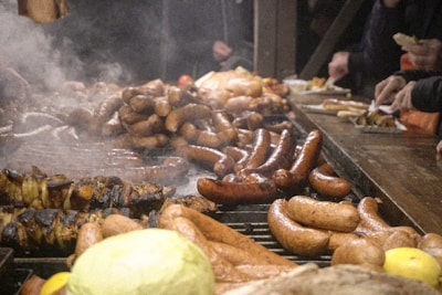 A variety of grilled sausages and meats displayed on a large barbecue grill, with steam rising from the cooked items. Several people are reaching for the food, suggesting a food market or gathering. A mix of cooked vegetables, such as peppers and onions, are also visible.