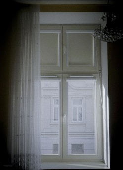 A window with sheer curtains partially covering a view of an exterior building, featuring architectural details and multiple windows. A chandelier hangs at the upper right corner, adding a touch of elegance.