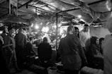 A bustling market scene with people gathered under fabric-covered stalls. Several individuals are seated and standing around a table beneath bright lights. There is a sense of lively interaction as vendors and customers engage with each other.