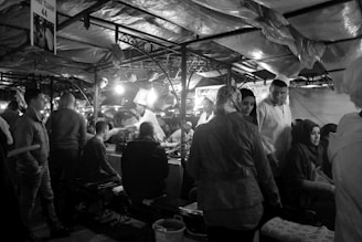 A bustling market scene with people gathered under fabric-covered stalls. Several individuals are seated and standing around a table beneath bright lights. There is a sense of lively interaction as vendors and customers engage with each other.