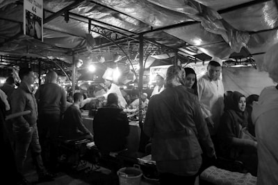 A bustling market scene with people gathered under fabric-covered stalls. Several individuals are seated and standing around a table beneath bright lights. There is a sense of lively interaction as vendors and customers engage with each other.
