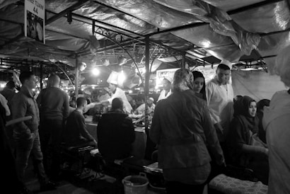 A bustling market scene with people gathered under fabric-covered stalls. Several individuals are seated and standing around a table beneath bright lights. There is a sense of lively interaction as vendors and customers engage with each other.