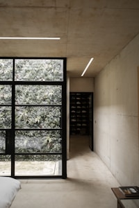 A minimalist interior features a large window providing a view of lush greenery outside, contrasting against the smooth, light-colored concrete walls. There's a corridor leading to a space filled with shelves, possibly a closet or storage area. A small table holds a book or magazine, and part of a bed is visible.