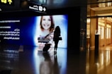 A silhouette of a person pulling a suitcase is seen in front of a large digital screen displaying an advertisement featuring a smiling child holding a model of a city landscape. The screen includes text in both English and Arabic. Above, signs point towards a restaurant, with reflections visible on the shiny floor.