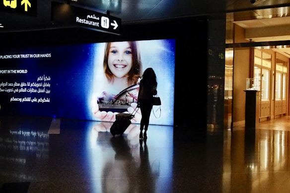 A silhouette of a person pulling a suitcase is seen in front of a large digital screen displaying an advertisement featuring a smiling child holding a model of a city landscape. The screen includes text in both English and Arabic. Above, signs point towards a restaurant, with reflections visible on the shiny floor.