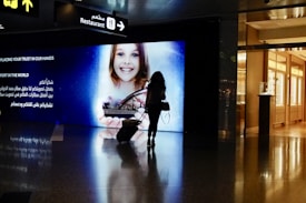 A silhouette of a person pulling a suitcase is seen in front of a large digital screen displaying an advertisement featuring a smiling child holding a model of a city landscape. The screen includes text in both English and Arabic. Above, signs point towards a restaurant, with reflections visible on the shiny floor.