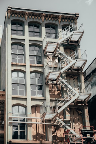 A multi-story building featuring large arched windows and a weathered exterior. A prominent metal fire escape zigzags down its side, casting intricate shadows on the facade. The overall look is aged, with signs of renovation visible through scaffolding and protective coverings.