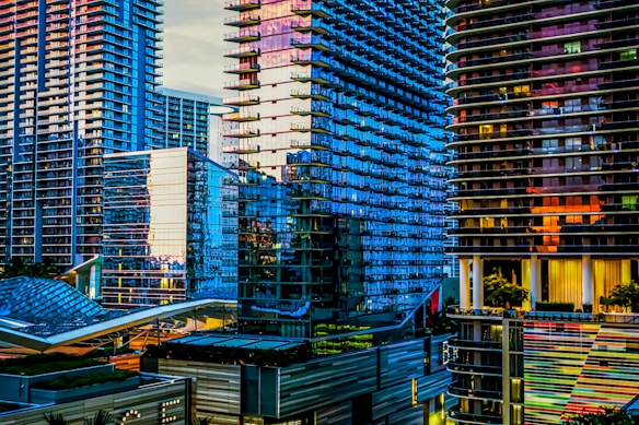 A vibrant cityscape featuring modern skyscrapers with reflective glass facades, illuminated by sunlight creating an array of colors. The scene showcases architectural elements with emphasis on linear designs and patterns.