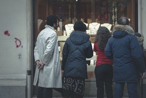 Several people stand outside a jewelry store, peering into a brightly lit display of necklaces and other jewelry items. The group includes individuals dressed in winter attire, such as coats and jackets. The setting suggests a busy urban street.