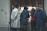 Several people stand outside a jewelry store, peering into a brightly lit display of necklaces and other jewelry items. The group includes individuals dressed in winter attire, such as coats and jackets. The setting suggests a busy urban street.
