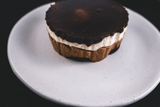 A dessert with layers of chocolate and cream sits on a white plate. The top layer is dark chocolate, with a creamy middle layer and a base that appears to be a chocolate crust. The plate is plain and white, contrasting with the rich colors of the dessert.