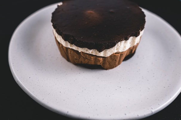 A dessert with layers of chocolate and cream sits on a white plate. The top layer is dark chocolate, with a creamy middle layer and a base that appears to be a chocolate crust. The plate is plain and white, contrasting with the rich colors of the dessert.