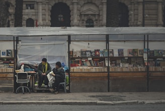 A street bookstall is set up with a variety of books displayed on wooden shelves. Two people are sitting under the shelter, engaged in conversation or reading. The background shows an old, stately building with arches and pilasters.