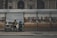 A street bookstall is set up with a variety of books displayed on wooden shelves. Two people are sitting under the shelter, engaged in conversation or reading. The background shows an old, stately building with arches and pilasters.