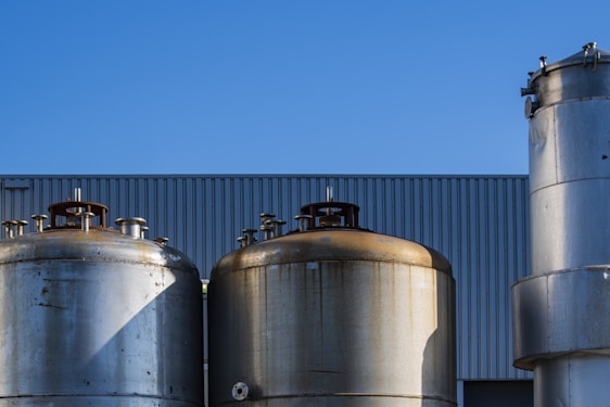 Several large industrial storage tanks are set against a backdrop of a clear blue sky. The tanks are metallic and have a weathered appearance, with some showing signs of rust. They are positioned in front of a building with a corrugated metal facade.