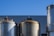 Several large industrial storage tanks are set against a backdrop of a clear blue sky. The tanks are metallic and have a weathered appearance, with some showing signs of rust. They are positioned in front of a building with a corrugated metal facade.