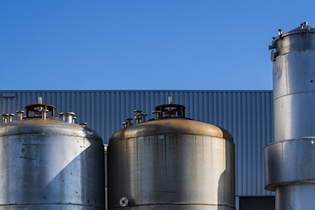 Several large industrial storage tanks are set against a backdrop of a clear blue sky. The tanks are metallic and have a weathered appearance, with some showing signs of rust. They are positioned in front of a building with a corrugated metal facade.